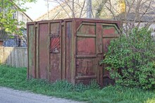 One Old Square Brown Red Rusty Container With A Window Behind Bars Stands In Green Grass And Vegetation On The Street