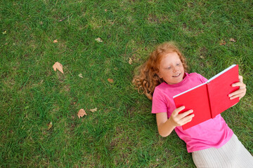 Poster - Little redhead girl reading book on green grass