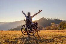 Woman Sitting On A Wheelchair Enjoying Sundown On Mountain Background.