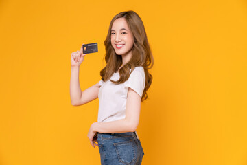 Wall Mural - Cheerful beautiful Asian woman in white t-shirt and holding mockup credit card on yellow background.