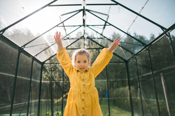 Wall Mural - Happy little girl standing with hands raised in eco greenhouse and looking at camera.