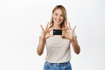 Wall Mural - Smiling young blond woman showing credit card, advertisement of bank, contactless payment, standing in t-shirt over white background