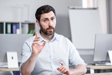 Young handsome successful business coach explains and tells on camera, gestures with his hands. Sitting at a computer desk in the office, looking at the camera