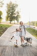 Cute woman mom hipster with tattoo rides skateboard and holding hands with daughter child and dog of Australian Shepherd breed sidewalk in park, warm summer evening and family time and hobbies