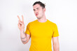© Roquillo - Young caucasian man wearing yellow t-shirt over white background makes peace gesture keeps lips folded shows v sign. Body language concept