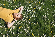 Young Man Laying In Green Grass With Flowers. People Fatigue From Work. Summer Sleeping And Relaxation Techniques. Vitamin D Sunbathing. Man Power Nap With Eye Closed. Rest After Work From Home