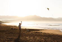 Young Boy Flying Kite At Sunrise On The Shore Of The Beach