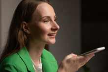 Young Woman Calls To Friend Via Mobile Phone Telling About First Working Day. Brunette Lady In Green Jacket Holds Device On Blurred Background Closeup