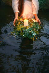 Women's hands close-up hold herbal ritual spiritual wreath, lay put float on water. Bright vintage burning candle, orange light. River, lake water, night summer nature. Slavic holiday Ivan Kupala