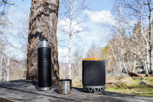 Tourist Utensils Stand On A Wooden One With A Tole In The Forest, A Camp Kitchen, Lunch In Nature, A Thermos With Tea, A Pan With Food, A Mug With Coffee.