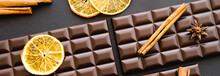 Top View Of Cinnamon And Dry Orange Sticks On Chocolate Bars On Black Background, Banner.