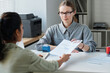 © AnnaStills - Mature Caucasian female consular officer sitting at desk in front of applicant for travel visa giving approval after successful interview