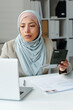 © AnnaStills - Vertical portrait of young adult Muslim woman wearing hijab sitting at desk in consulate office working with documents using laptop