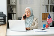 © AnnaStills - Portrait of young adult Muslim woman wearing hijab working as consular officer in US embassy texting on laptop and talking on phone
