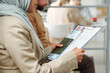 © AnnaStills - Unrecognizable Muslim woman wearing hijab sitting on chair waiting for appointment in queue holding documents for visa application