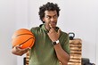 African man with curly hair holding basketball ball at the gym serious face thinking about question with hand on chin, thoughtful about confusing idea