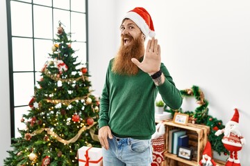 Poster - Redhead man with long beard wearing christmas hat by christmas tree waiving saying hello happy and smiling, friendly welcome gesture