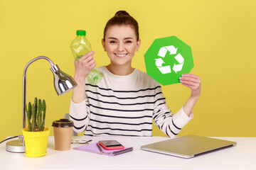 Portrait of satisfied woman with charming smile holding empty plastic bottle and green recycling sign, sitting on workplace. Indoor studio studio shot isolated on yellow background.