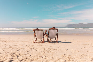 African american couple holding hands while relaxing on deckchairs at beach against sky in summer