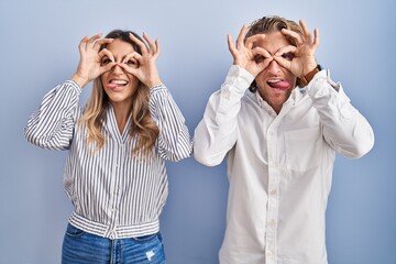 Poster - Young couple standing over blue background doing ok gesture like binoculars sticking tongue out, eyes looking through fingers. crazy expression.