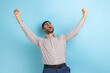 © khosrork - Portrait of of overjoyed handsome businessman expressing winning gesture with raised fists and screaming, celebrating victory, wearing striped shirt. Indoor studio shot isolated on blue background.