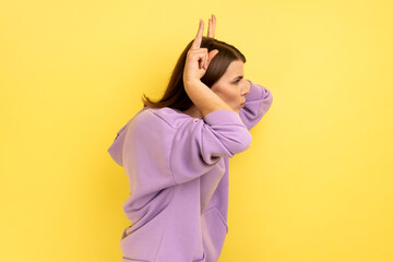 Side view of aggressive woman holding fingers above head showing horns, arrogant and stubborn, ready to attack, wearing purple hoodie. Indoor studio shot isolated on yellow background.