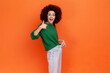 © khosrork - Portrait of positive excited woman with Afro hairstyle wearing green casual style sweater showing slim waist in big trousers and thumbs up. Indoor studio shot isolated on orange background.