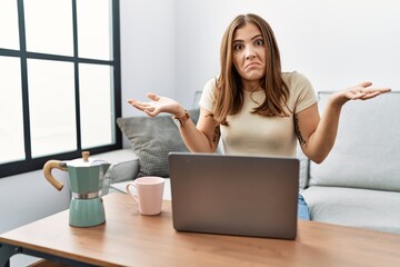 Sticker - Young brunette woman using laptop at home drinking a cup of coffee clueless and confused expression with arms and hands raised. doubt concept.