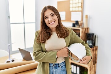 Canvas Print - Young brunette woman holding architect hardhat at the office looking positive and happy standing and smiling with a confident smile showing teeth