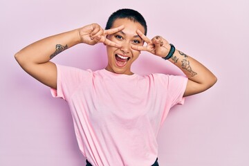 Wall Mural - Beautiful hispanic woman with short hair wearing casual pink t shirt doing peace symbol with fingers over face, smiling cheerful showing victory