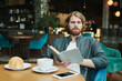 © Mediaphotos - Confident young man in casual shirt sitting in armchair and looking away while talking by phone in modern cafe