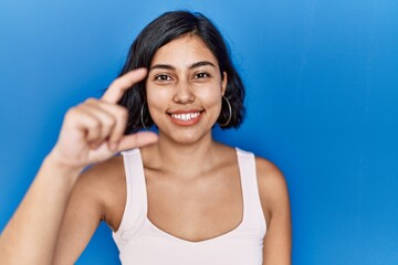 Canvas Print - Young hispanic woman standing over blue background smiling and confident gesturing with hand doing small size sign with fingers looking and the camera. measure concept.