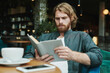 © Mediaphotos - Serious handsome guy with beard sitting at table in cafe and drinking coffee while reading mail on smartphone
