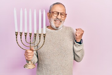 Sticker - Handsome senior man with beard holding menorah hanukkah jewish candle screaming proud, celebrating victory and success very excited with raised arm