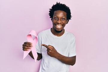 Sticker - Young african american man holding pink cancer ribbon smiling happy pointing with hand and finger