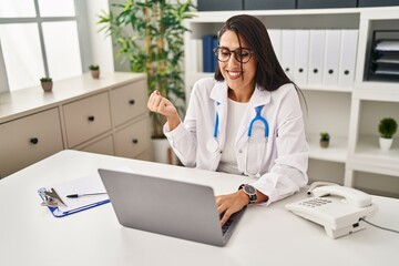 Wall Mural - Young hispanic doctor woman doing video call at the clinic screaming proud, celebrating victory and success very excited with raised arm