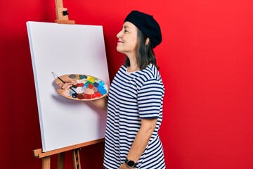 Poster - Middle age hispanic woman standing drawing with palette by painter easel stand looking to side, relax profile pose with natural face and confident smile.