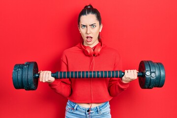 Poster - Beautiful woman with blue eyes wearing sportswear using dumbbells in shock face, looking skeptical and sarcastic, surprised with open mouth