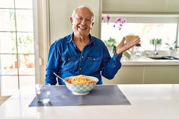 Wall Mural - Senior man with grey hair eating pasta spaghetti at home smiling cheerful presenting and pointing with palm of hand looking at the camera.