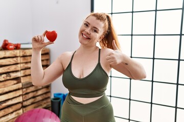 Canvas Print - Young redhead woman holding heart at the gym smiling happy and positive, thumb up doing excellent and approval sign