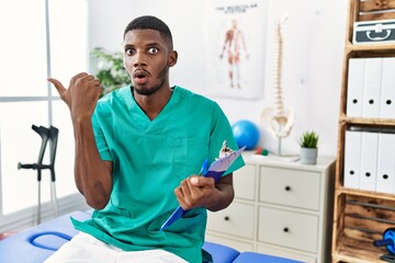 Canvas Print - Young african american man working at pain recovery clinic surprised pointing with hand finger to the side, open mouth amazed expression.