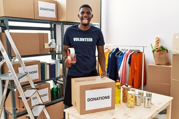 Poster - Young african american volunteer man packing donations box for charity sticking tongue out happy with funny expression. emotion concept.