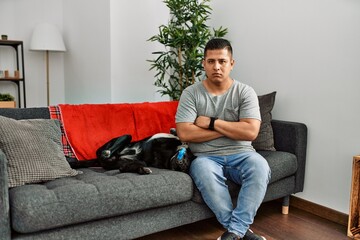 Poster - Young latin man and dog sitting on the sofa at home skeptic and nervous, disapproving expression on face with crossed arms. negative person.