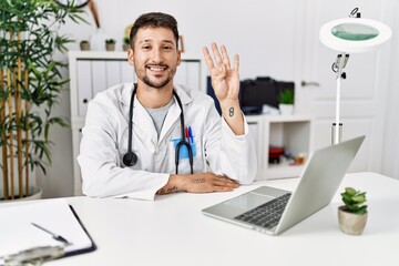 Poster - Young doctor working at the clinic using computer laptop showing and pointing up with fingers number four while smiling confident and happy.