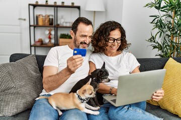 Wall Mural - Middle age hispanic couple using laptop and credit card. Sitting on the sofa with dogs at home.