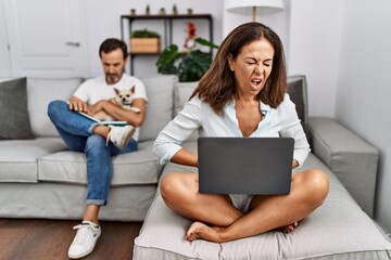 Sticker - Hispanic middle age couple at home, woman using laptop with hand on stomach because nausea, painful disease feeling unwell. ache concept.