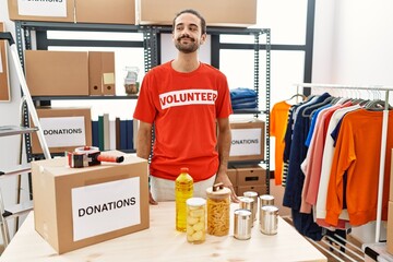 Wall Mural - Young hispanic man wearing volunteer t shirt at donations stand smiling looking to the side and staring away thinking.