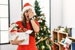 Young brunette woman standing by christmas tree holding decoration yawning tired covering half face, eye and mouth with hand. face hurts in pain.
