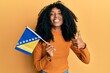 African american woman with afro hair holding bosnia herzegovina flag smiling happy and positive, thumb up doing excellent and approval sign