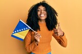 Fototapeta  - African american woman with afro hair holding bosnia herzegovina flag smiling happy and positive, thumb up doing excellent and approval sign